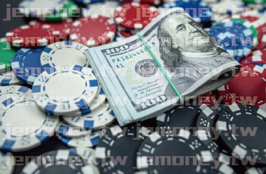 「畫面中央為一疊綁橡皮筋的百元美鈔,周圍散落紅藍白黑四色籌碼。」